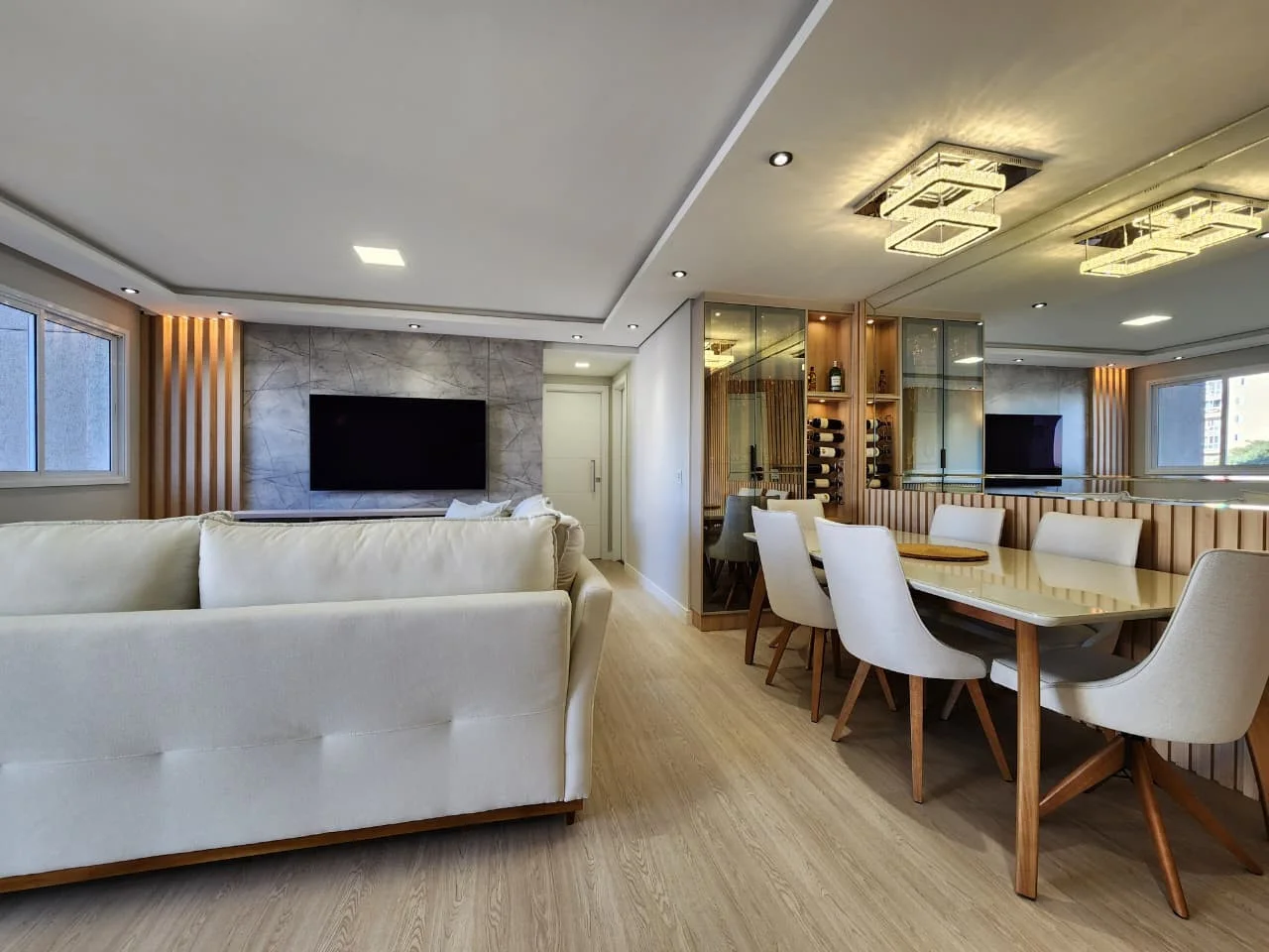 Living e sala de jantar integrados, com painel de TV em porcelanato marmorizado, adega climatizada em vidro e boiserie ripada atrás da mesa